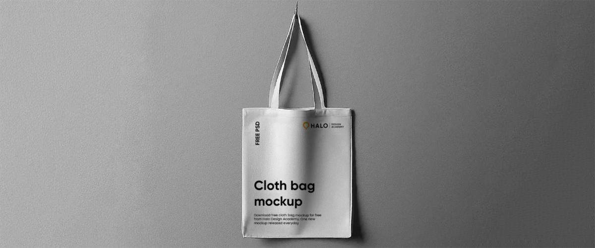 A white cotton tote bag mockup hanging on a gray wall for free cloth bag design freebie