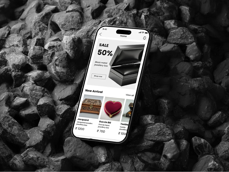 A smartphone mockup of a jewelry box e-commerce app resting on a bed of dark, textured coal rocks
